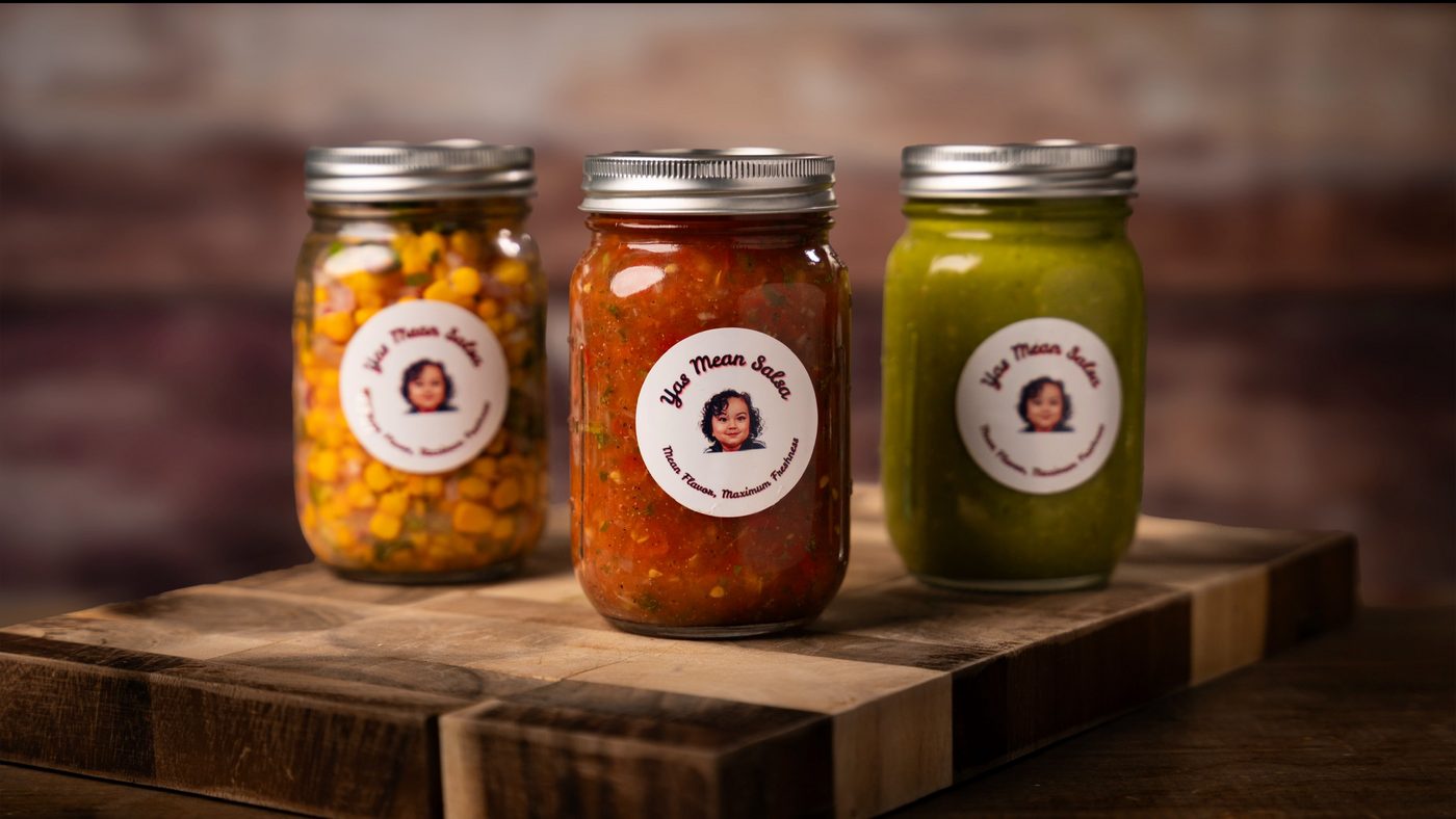 Three labeled jars of Yas Mean Salsa — corn, red, and green — lined up on a wooden board