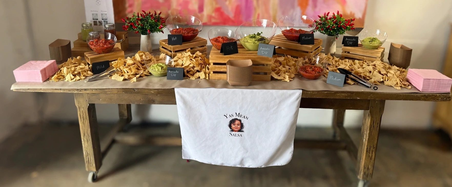 Yas Mean Salsa Bar setup at an event — bowls of Mild, Hot, and Avocado Dip with tortilla chips spread across a wooden table under the Yas Mean Salsa banner